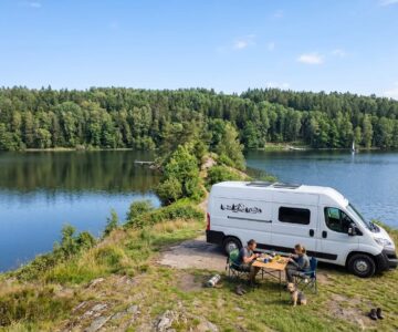 Furgoneta camper junto a un lago con dos personas comiendo al aire libre y un perro.
