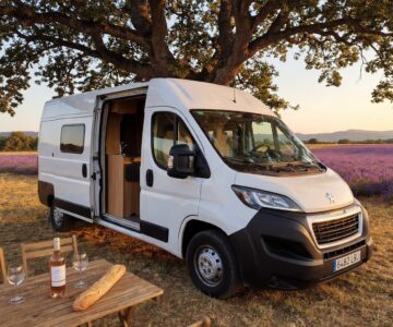 Furgoneta camper blanca junto a un campo de lavanda con una mesa con vino y pan.