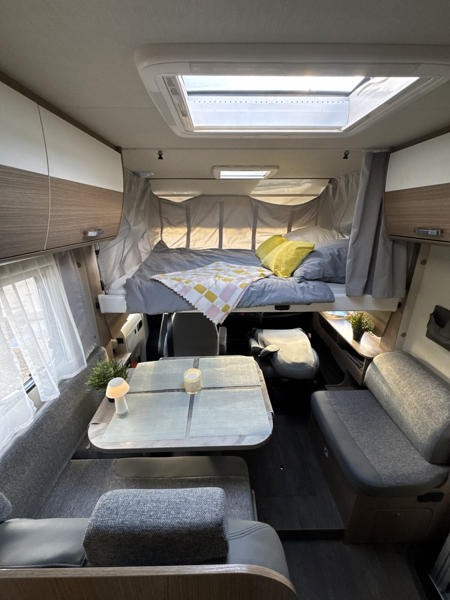 Cama abatible sobre cabina en autocaravana con ropa de cama completa, armarios laterales y zona comedor inferior preparada